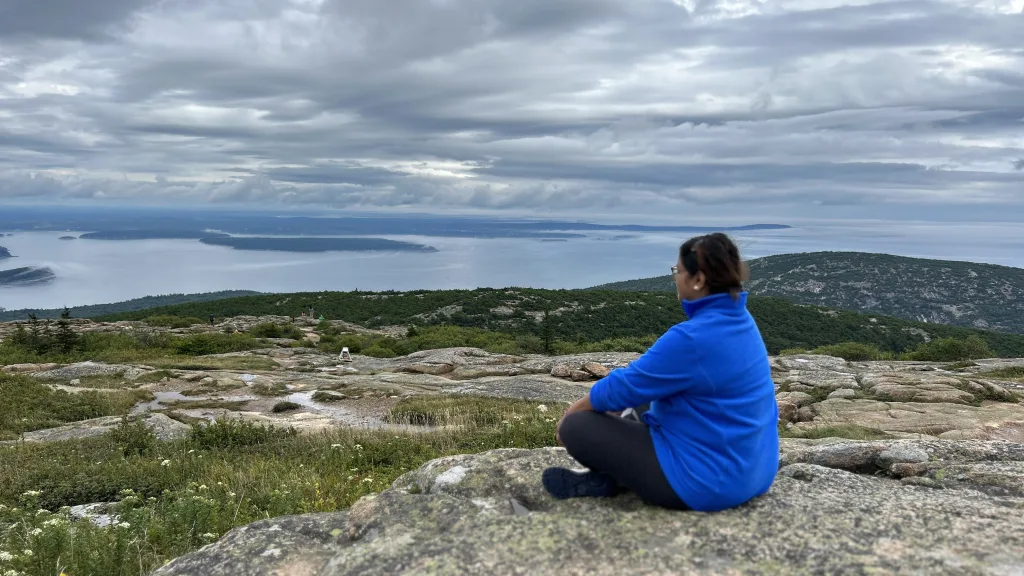 A woman sitting cross legged on the ground looking out over the waters in Acadia